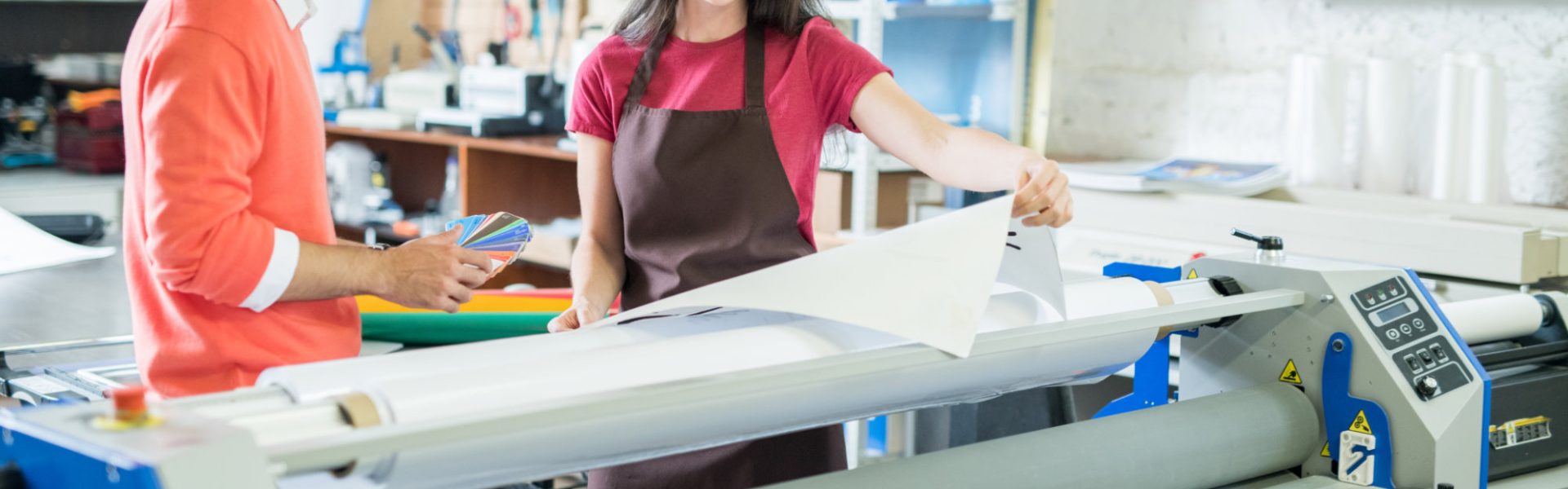 Cheerful excited beautiful lady in apron showing printed banner to manager who picking out color on swatch for printing